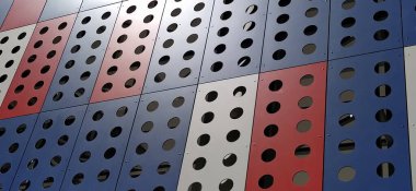 Modern bir binanın cephesi. Kırmızı, mavi ve beyaz levhalar ya da dekoratif delikli metal profiller. Dış yüzeyde teknolojik çözümler. Dekoratif elementler altında destekleyici yapılar inşa ediliyor.