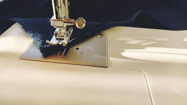 Sewing process on a modern sewing machine, close-up. Sewing machine foot with a needle. Dark blue fabric is trimmed with double wrapping of the fabric edge. Natural sunlight.