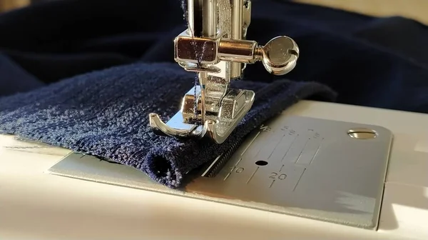 Sewing process on a modern sewing machine, close-up. Sewing machine foot with a needle. Dark blue fabric is trimmed with double wrapping of the fabric edge. Natural sunlight.