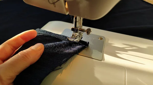 Sewing process on a modern sewing machine, close-up. Sewing machine foot with a needle. Dark blue fabric is trimmed with double wrapping of the fabric edge. Natural sunlight.