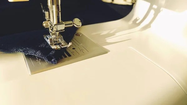 Sewing process on a modern sewing machine, close-up. Sewing machine foot with a needle. Dark blue fabric is trimmed with double wrapping of the fabric edge. Natural sunlight.