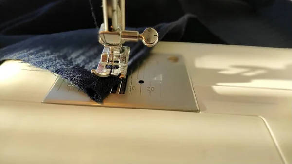 Sewing process on a modern sewing machine, close-up. Sewing machine foot with a needle. Dark blue fabric is trimmed with double wrapping of the fabric edge. Natural sunlight.