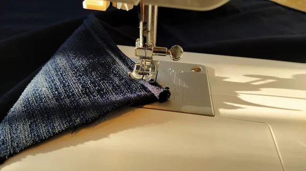 Sewing process on a modern sewing machine, close-up. Sewing machine foot with a needle. Dark blue fabric is trimmed with double wrapping of the fabric edge. Natural sunlight.