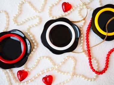 Düz yatıyordu. Cam siyah tabaklardan, plastik mücevherlerden, bileziklerden, boncuklardan, beyaz arka planda kırmızı kalplerden oluşan güzel bir kompozisyon. 14 Şubat Sevgililer Günü için kadın takıları. Durgun hayat.