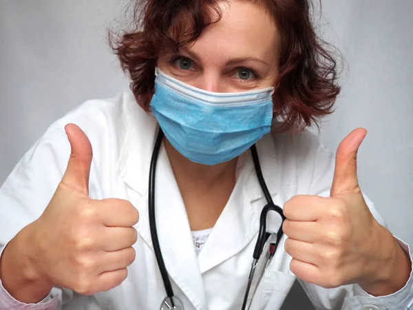 Doctor shows with hands a thumb up gesture. Cheerful look. A medical professional in a white coat and with a stethoscope. A woman doctor in a surgical mask looks at you and smiles under the mask
