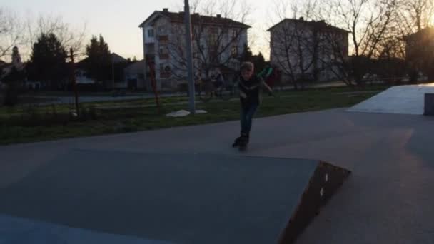 Een jongen van 8 jaar oud rolschaatst op het sportveld. Het kind rolt op een bult op rollen en rolt naar beneden aan de andere kant. Buitenactiviteiten of training. Jongen met blond haar en grijze ogen — Stockvideo