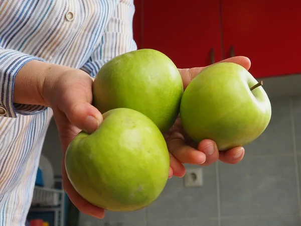 Maçãs na mão. A mulher segura três maçãs verdes em suas mãos e as oferece ao espectador. Comida orgânica. Frutas frescas para comida. Grande maçã azeda. Camisa branca com riscas. Cozinha com mobiliário vermelho — Fotografia de Stock