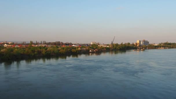 Banco del río con una zona industrial. Flujo de agua en el río Sava, Serbia, Sremska Mitrovica, Balcanes. Se enfría en la superficie del agua. Hora dorada. Edificios, estructuras de fábrica y barcazas — Vídeo de stock