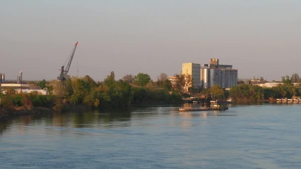 Pråmen avgår från flodstranden med en industrizon. Vattenflöde på floden Sava, Serbien, Sremska Mitrovica. Ta det lugnt på vattenytan. Byggnader, fabriksbyggnader, pråmar, hamnar. — Stockvideo