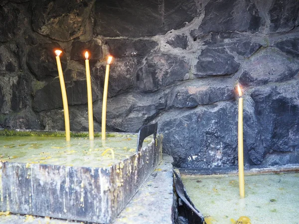 Burning candles for peace or health. Church religious rite. Three candles on the left on the stand, one candle on the right. Melted wax in a metal container. Stone background.