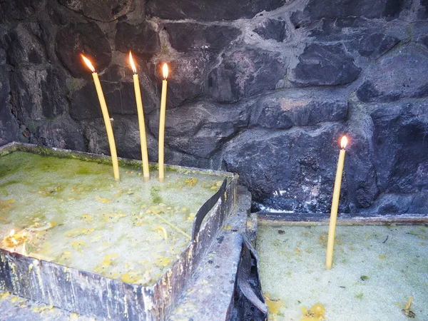 Burning candles for peace or health. Church religious rite. Three candles on the left on the stand, one candle on the right. Melted wax in a metal container. Stone background