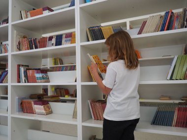 Sremska Mitrovica, Sırbistan 15 Mayıs 2021. 7 yaşında beyaz tişörtlü bir kız kütüphanedeki kitaplığın yanında duruyor ve kitap okuyor. Okul eğitimi. Edebiyat ve okuma aşkı.