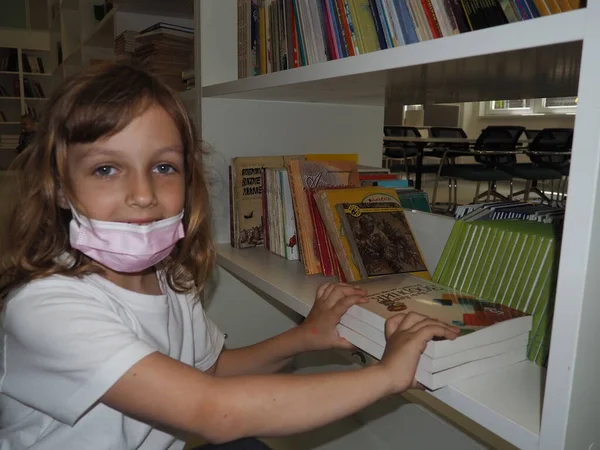 Sremska Mitrovica Servia 2021年5月15日図書館の本棚の近くにマスクと白いTシャツを着た7歳の少女が立っていて 手で本に触れています 学校教育 — ストック写真