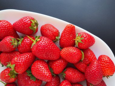 Tabakta çilek, siyah arka plan. Beyaz tabaklar ve lezzetli kırmızı büyük bahçe çilekleri. Sağlıklı yiyecekler, vitaminler ve vejetaryen diyeti. Yiyeceklerle yaşam. Uzayı kopyala Düz yatıyordu.