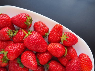 Tabakta çilek, siyah arka plan. Beyaz tabaklar ve lezzetli kırmızı büyük bahçe çilekleri. Sağlıklı yiyecekler, vitaminler ve vejetaryen diyeti. Yiyeceklerle yaşam. Uzayı kopyala Düz yatıyordu.