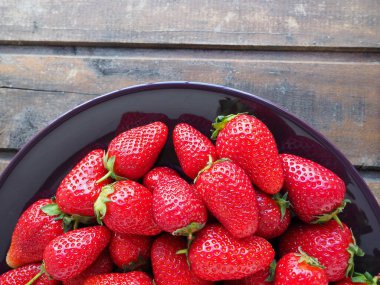 Tabakta çilek, ahşap arka plan. Koyu seramik tabaklar ve lezzetli kırmızı büyük bahçe çilekleri. Sağlıklı yiyecekler, vitaminler ve vejetaryen diyeti. Yiyeceklerle yaşam. Uzayı kopyala Düz yatıyordu