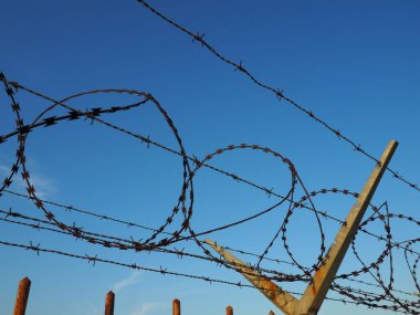 Mavi gökyüzüne uzanan dikenli teller. Hırsızlara ve vandallara karşı dikenli tel. Üstesinden gelinemez bir engel. Paslı tel, sıralar, spiral ve dikenli tel bobinler