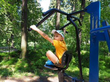 8 yaşındaki sarışın bir çocuk spor aletleri veya simülatörle ormanda vücut geliştirme sporu yapıyor. El ve sırt kasları. Çocuk egzersiz sırasında etrafına bakar..