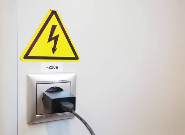 High-voltage sign on the white wall. A plug with a cable is stuck in an electrical outlet. Charging your phone or smartphone. The danger of electric shock. Black arrow on a yellow background