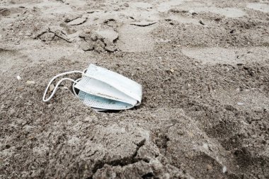 Kullanılmış bir tıbbi maske kumlu bir plaja bırakılmış. Kumun üzerinde bir mavi yüz kalkanı var. Salgın koronavirüs covid-19. Tek kullanımlık ürünlerle ekoloji, geri dönüşüm ve çevre kirliliği.