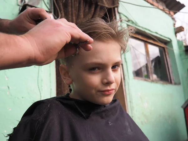 8歳の金髪の少年がはさみで髪を切る。自宅で美容師サービス。金髪の子供のためのヘアカット. — ストック写真
