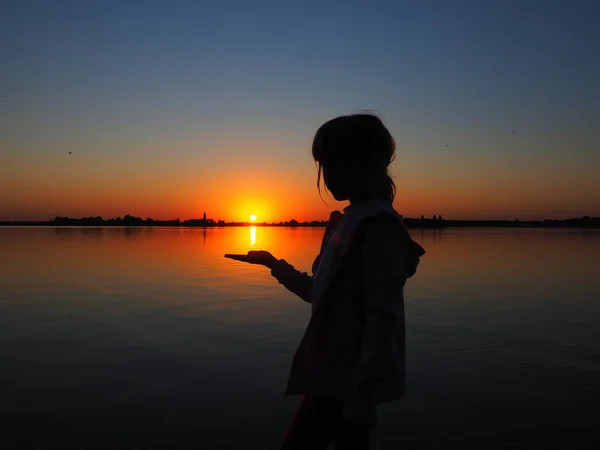 Silhouette d'une jeune fille sur fond de coucher de soleil. L'enfant tient le soleil couchant sur sa main. Horizon, eau, soleil et ciel en arrière-plan. Figurine noire vêtue de capuche. Lac Palic, Serbie — Photo