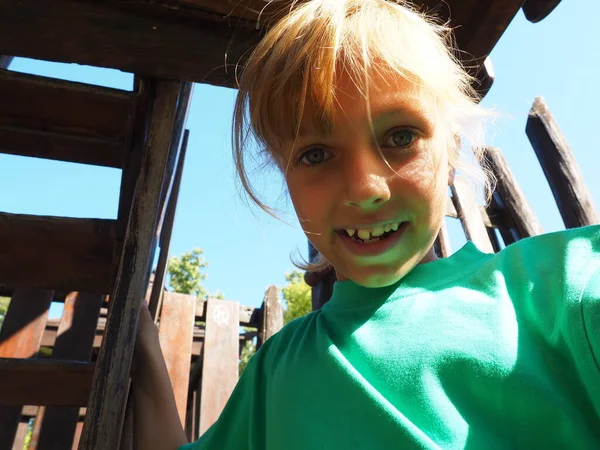 Una chica caucásica de 7 años con el pelo rubio juega en un parque infantil de madera y se ríe. Camiseta verde, pelo atado en una cola de caballo. Ausencia de un denículo lácteo de la mandíbula superior. Niño feliz camina en el sol —  Fotos de Stock