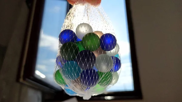 Multi-colored clickers or glass balls for decoration. Blue, green, beige balls are suspended in a net in the air against the window and the sky. The light of the sun shines through glass — Stock Photo, Image