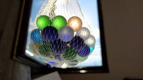 多种颜色的小丑或玻璃球用于装饰。蓝色、绿色、米黄色的球悬挂在空中的网中，靠着窗户和天空。阳光透过玻璃射出光芒 — 图库照片