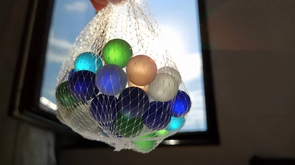 Clickers multicoloridos ou bolas de vidro para decoração. Bolas azuis, verdes e bege são suspensas em uma rede no ar contra a janela e o céu. A luz do sol brilha através do vidro — Fotografia de Stock