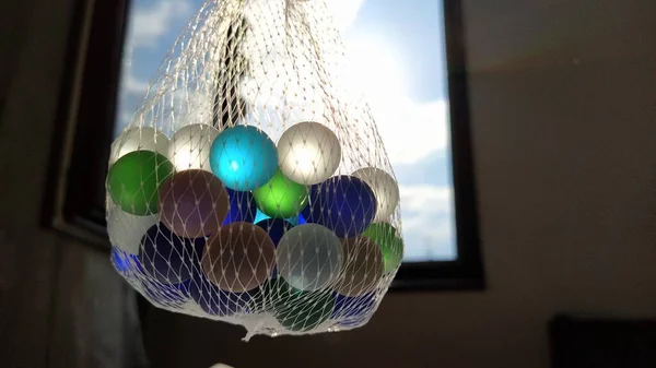 Multi-gekleurde klikkers of glazen ballen voor decoratie. Blauwe, groene, beige ballen hangen in een net in de lucht tegen het raam en de lucht. Het licht van de zon schijnt door glas — Stockfoto