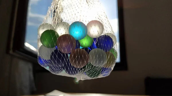 Πολύχρωμα clickers ή γυάλινες μπάλες για διακόσμηση. Μπλε, πράσινες, μπεζ μπάλες αιωρούνται σε ένα δίχτυ στον αέρα ενάντια στο παράθυρο και τον ουρανό. Το φως του ήλιου λάμπει μέσα από το γυαλί — Φωτογραφία Αρχείου
