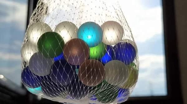 Multi-gekleurde klikkers of glazen ballen voor decoratie. Blauwe, groene, beige ballen hangen in een net in de lucht tegen het raam en de lucht. Het licht van de zon schijnt door glas — Stockfoto
