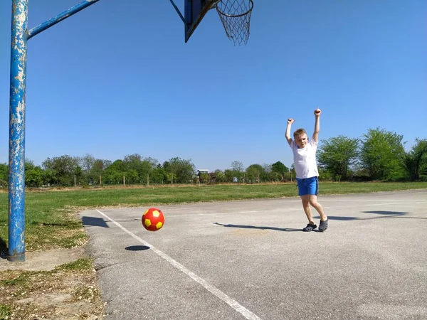 Sremska Mitrovica, Sırbistan. 6 Haziran 2020. Bir çocuk oyun alanında top oynuyor. Asfalt spor sahası. Beyaz tişörtlü bir çocuk. Sarı saçlı, 7 yaşında yeni yürümeye başlamış bir çocuk. Koşmak, tekmelemek ve egzersiz yapmak — Stok fotoğraf