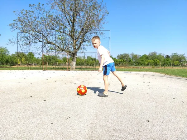 Sremska Mitrovica, Serbien. 6 juni 2020. En pojke spelar boll på lekplatsen. Asfalt idrottsplan. Ett barn i vit t-shirt. Småbarn med blont hår, sju år. Löpning, spark och övningar — Stockfoto