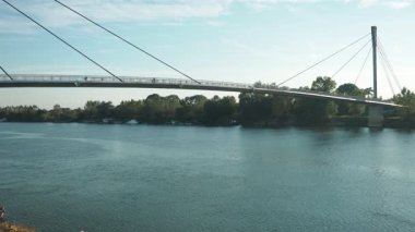 Sremska Mitrovica, Sırbistan, 17 Ekim 2025: Barışçıl Sava nehrinin yüzeyinde parlak güneş ışığı parlıyor. Modern köprü su üzerinde kavis çizer, dingin bir atmosfer yaratır. Yemyeşil ağaçlar kıyılarda.