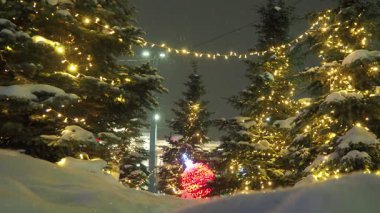 Şehir meydanının Noel süsü. Kar yağışlı kış akşamı. Noel ağaçları parlak ampuller ve çelenklerle süslenir. Karda, Noel balosu şeklinde büyük kırmızı bir lamba. Yeni Yıl Manzarası.