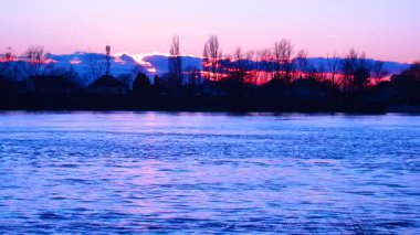 Gökyüzü renkleri değiştirirken güneş nehrin üzerinde batar. Yansımalar suda dans eder. Ağaçlar kıyı boyunca hareketsiz duruyor. Gün, doğanın bir gösterisiyle sona erer..