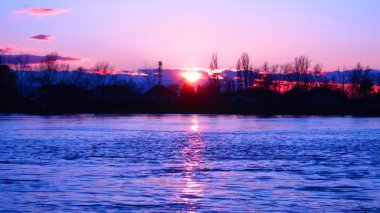 Gün sona ererken, Sırbistan 'daki barış dolu Sava Nehri üzerinde güneş batıyor. Altın ışık su yüzeyinde dans ederken yumuşak pembe renkler mavi gökyüzünü doldurarak büyülü bir ortam yaratır.