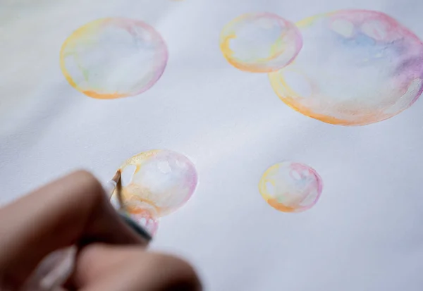 Female hand coloring bubble drawing with colored watercolors in a sketchbook on a white table.