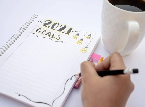 Decorated notebook for writing New Year\'s resolutions on a white table with a black coffee cup on the side and a hand writing with a black pencil.