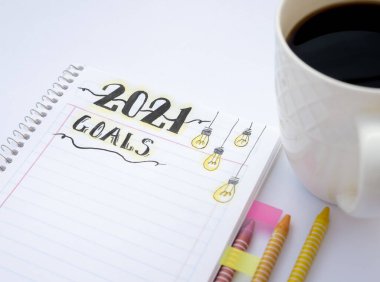 Renkli boya kalemleri ve yanında siyah kahve fincanı olan beyaz bir masanın kenarlarına renkli ayırıcılarla yeni yıl kararları yazmak için dekore edilmiş bir defter..
