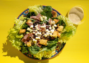 Ekmek arası, marul, domates, jambon, ton balığı, peynir ve meyveli nefis sağlıklı salata. Sarı bir masanın üzerinde el yapımı bir tabakta..