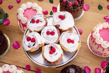 Gümüş tepside kırmızı böğürtlenlerle kaplı nefis tarçın rolleri ve etrafındaki Sevgililer Günü pastaları gül yapraklarıyla süslenmiş ahşap bir masada..