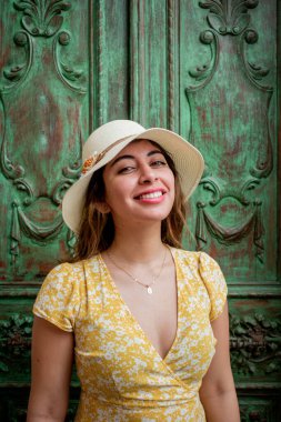 Renkli sarı elbisesi ve beyaz şapkasıyla gülümseyen güzel bir genç kadın Mexico City 'de eski bir koloni tarzı yeşil kapının önünde..