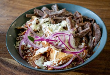 Lezzetli geleneksel Meksika kırmızı Chilaquiles peynirli, kırmızı soğanlı, kremalı ve biftekli ahşap bir tabakta.