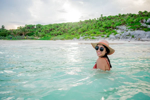 Beautiful woman smiling in an orange swimsuit, white hat and black glasses in the blue sea of Quintana Roo in Mexico.