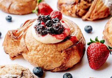 Üzerinde böğürtlen ve çilek olan peynirli kruvasan hamuru beyaz bir masada ve etraftaki tatların rolleri yakın konseptte..