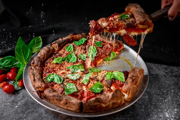 Appetizing Chicago-style pizza with American salami, Italian sausage, albacore and mozzarella cheese with a slice held by a spatula with tomatoes and baby spinach in the background
