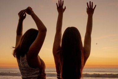 Sahilde dans eden Latin kızlar, arka planda günbatımı var.